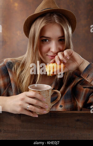 Young beautiful blonde in a brown plaid shirt with a mug of coffee and a cupcake .Natural old wooden background . Stock Photo