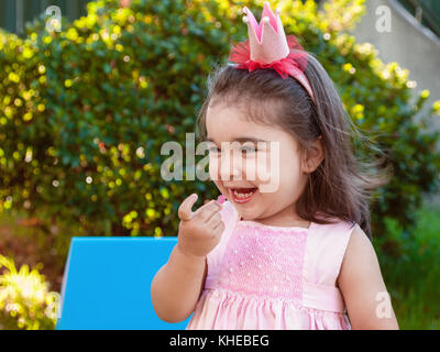 Very happy baby toddler girl, eating gummies laughing and smiling in outdoor party dressed in pink dress as princess or queen with crown Stock Photo