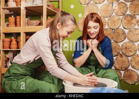 The female potter teaches a red-haired woman to properly mold a deep bowl of clay on a potter's wheel. Training potter to work on a potter's wheel, si Stock Photo
