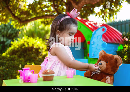 Baby toddler girl playing in outdoor tea party feeding her best friend Teddy Bear with candy gummy. Pink dress and queen or princess crown. Stock Photo