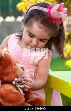 Baby toddler girl, playing in a tea party feeds best friend bff Teddy Bear with candy gummy. Pink dress and queen or princess crown Stock Photo