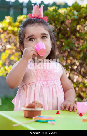 Baby toddler girl playing in outdoor tea party drinking from cup with lollipop, muffin and gummies on table. Pink dress and queen or princess crown Stock Photo