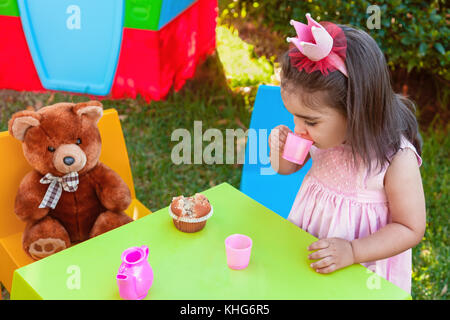 Baby toddler girl playing in outdoor tea party drinking from cup with best friend Teddy Bear sitting at table. Pink dress and queen or princess crown Stock Photo