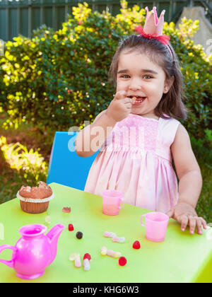 Happy baby toddler girl, eating gummies laughing and smiling in outdoor tea party dressed in pink dress as princess or queen with crown Stock Photo