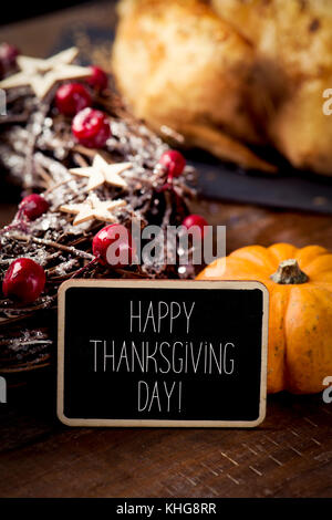 a black signboard with the text happy thanksgiving day written in it and a roast turkey and a pumpkin, placed on a rustic wooden table Stock Photo