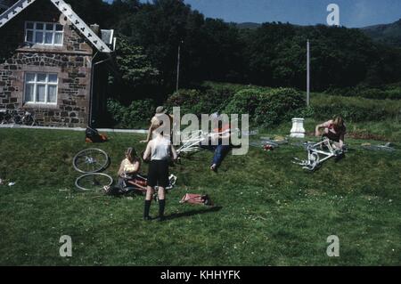 A photograph of five people on a short grass covered hill, four of the people are seated and are working on performing maintenance on bicycles, the fifth person stands watching the others, a section of a stone building can be seen in the background along with a field and trees, 1961. Stock Photo