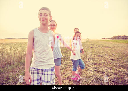 Four young caucasian kids holding hands, walking at cultivated countryside field, at summer sunset, wearing casual summer clothes Stock Photo