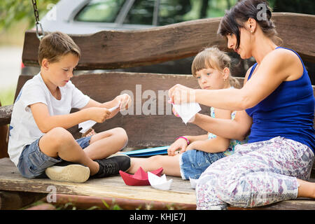 Caucasian three member family sitting outdoors on a big wooden swing. Mother is teaching her kids how to fold a paper boat Stock Photo