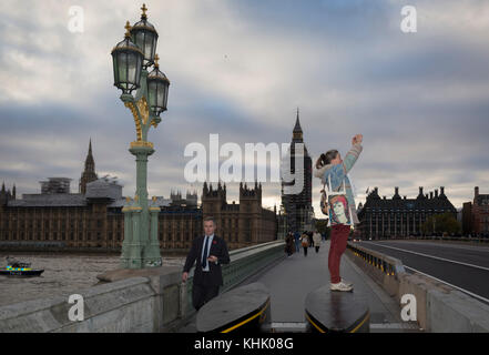 With the winter landscape of Westminster and the Houses of Parliament on the river Thames opposite, a woman with a David Bowie bag stands on the anti-terrorism security barriers at the southern end of Westminster Bridge, to take a picture of the London Eye, on 8th November 2017, in Lambeth, London. Stock Photo