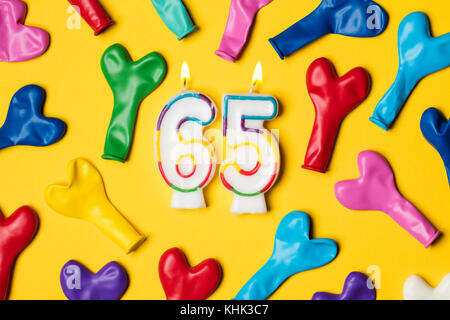 Number 65 candle with party balloons on a bright yellow background Stock Photo