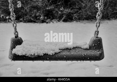 A swing sits with snow partly covering the seat, and beads of frozen ice sit on the seat edges,with the chrome chain covered in ice on a winters day. Stock Photo