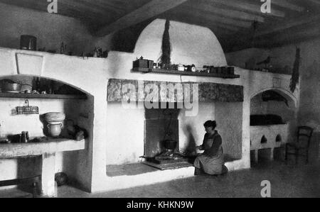 A photograph that shows a housewife cooking in the home of a herder, the large open fireplace where she is knelt down cooking is located centrally in the room, on the mantle are a number of kitchen items, to the left of the fireplace is an area that includes the washing basin and cooking utensils, Camargue, France, July, 1922. Stock Photo