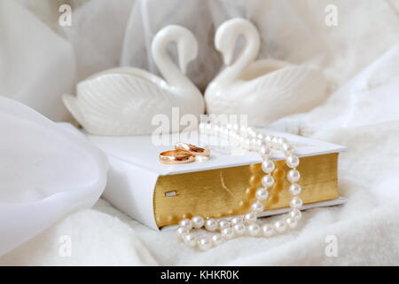 Two golden wedding rings on the white Bible. Also white pearls and two white porcelain swans. White background.