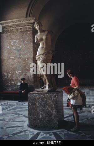 A photograph of the statue named Venus de Milo, the statue is believed to depict Aphrodite, a woman wearing a salmon colored top and carrying her coat and purse examines the statue from a close distance, a security guard sits on a bench in the background, the statue is located in the Louvre Museum, Paris, France, 1965. Stock Photo