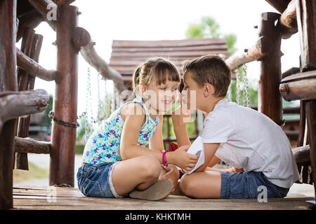 Happy two caucasian siblings sittign in a wooden house outdoors on summer day,  holding a paper boats in their hands, boy making psst gesture Stock Photo