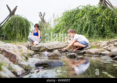 Two young caucasian kids laying on a small wooden bridge above small stream, dropping the boats to water Stock Photo