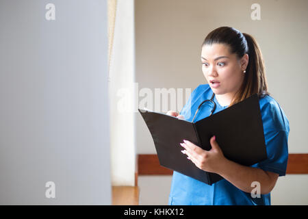 Young hispanic female doctor analyzing a clinical case, looking into a folder with surprised facial expression. Woman is wearing blue uniform, standin Stock Photo