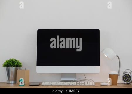 Electric lamp, mobile phone, desktop pc, disposable glass, flora and stationery on table against white wall Stock Photo