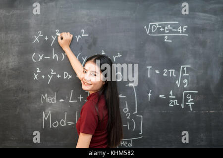 Beautiful Asian student writing on blackboard with chalk in classroom. Stock Photo