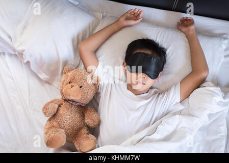 boy sleeping on bed with teddy bear white pillow and sheets wearing sleep mask.boy fall asleep in morning.sleep concept Stock Photo