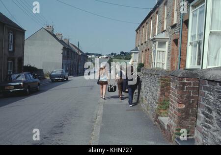 Three female travelers walking along a road beside old stone homes, two of the travelers hokding the two handles of a heavy suitcase and carrying it together, 1960. Stock Photo