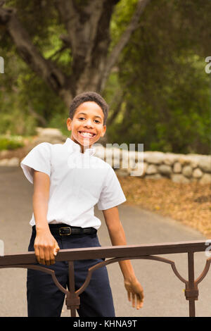 Happy African American boy standing, playing, or swinging on iron gate outside in front of trees at park. Space for copy in background area. Stock Photo