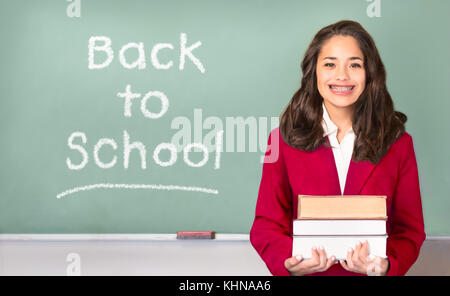 Back to School. Pretty ethnic or Hispanic teen with braces, wearing a red school uniform blazer isolated in front of green chalkboard with Back to sch Stock Photo