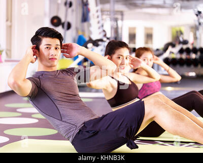 three young asian adults, male and female, doing sit-ups on the floor in gym. Stock Photo