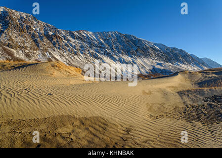 The picturesque mountain autumn landscape with snow covered mountains, frozen lake, sand dunes with sparse vegetation against the blue sky on a sunny  Stock Photo
