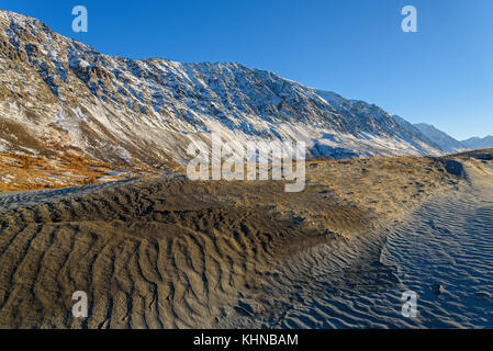 The picturesque mountain autumn landscape with snow covered mountains, frozen lake, sand dunes with sparse vegetation against the blue sky on a sunny  Stock Photo