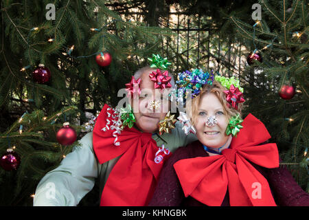 Couple covered in Christmas bows sitting under pine trees decorated with lights and red balls. Stock Photo