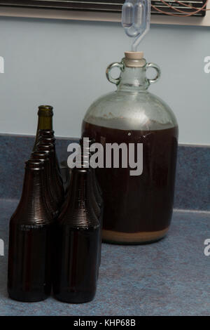 Fermented beer in a demijohn with airlock and dark brown bottles and over kitchen background Stock Photo