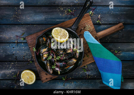 Delicious mussels. Serving on a hot frying pan with herbs spices and lemon on a colored wooden background. Top view. Stock Photo