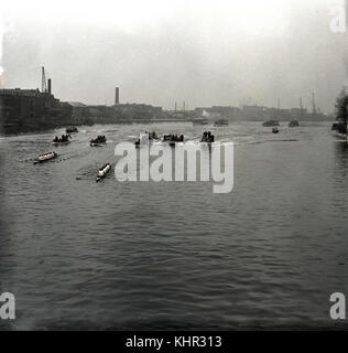 1950s, historial picture showing the boats and flotilla of the famous university rowing race, the Oxford & Cambridge Boat Race, going down the river Thames past the Hammersmith embankment, London, England. On the left, the many industrial wharves of this time, including the Manbre & Garton sugar warehouse. Stock Photo