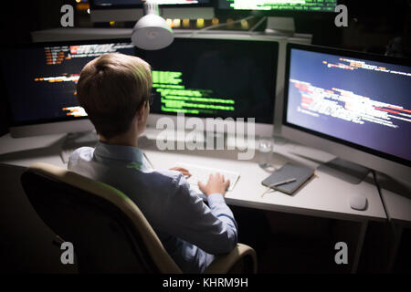 Back view of unrecognizable hacker stealing confidential data while sitting in front of modern computer, interior of dim room Stock Photo