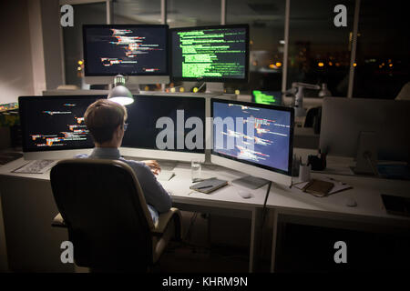 Back view of highly professional coder looking at wristwatch while sitting in front of computer and working on ambitious project, interior of dim open Stock Photo