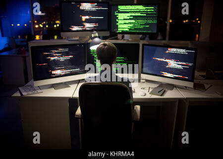 Rear view of hard-working programmer with stylish haircut sitting in front of computer and writing code, interior of dim open plan office on backgroun Stock Photo