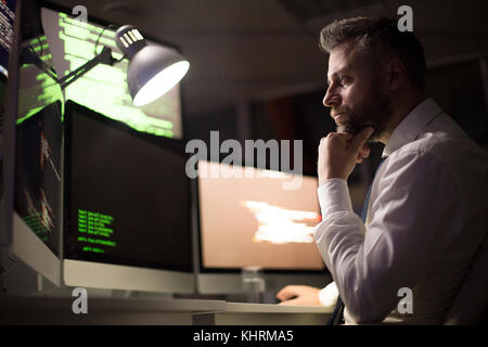 Profile view of pensive bearded coder wrapped up in work while sitting in front of modern computer, interior of dim office on background Stock Photo