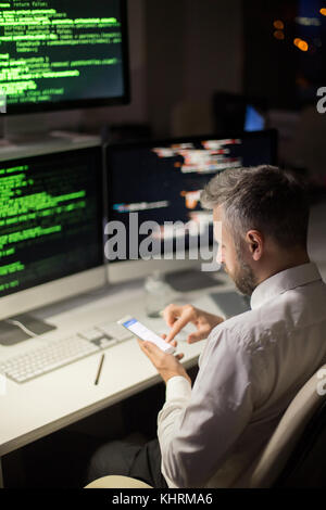 Confident middle-aged software developer texting with friend on smartphone while taking short break from work, interior of dim open plan office on bac Stock Photo