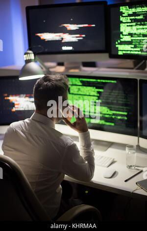 Back view of unrecognizable programmer wearing white shirt sitting in front of modern computer while talking to his colleague on smartphone, interior  Stock Photo
