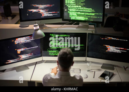 Unrecognizable gray-haired software developer writing code while sitting in front of modern computer, interior of dim office on background Stock Photo