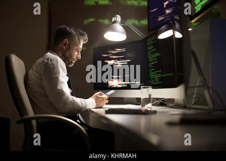 Profile view of talented middle-aged programmer using smartphone while sitting at office desk and finishing work on promising application, interior of Stock Photo