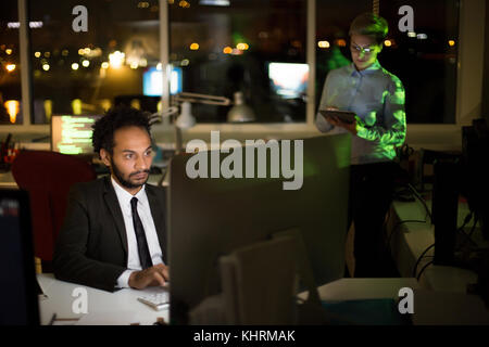 Busy dim open plan office: mixed race manager wearing stylish suit working on promising project with help of computer while his pretty young colleague Stock Photo