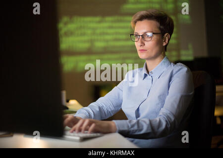 Pretty middle-aged entrepreneur sitting in front of computer and making necessary corrections in annual accounts, interior of dim office on background Stock Photo