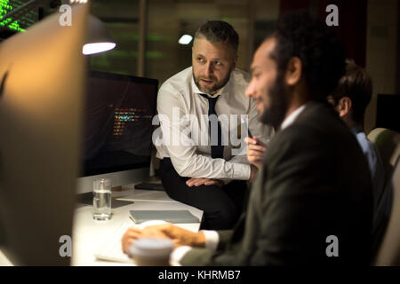 Team of software developers gathered together in front of modern computer and discussing details of their joint project, interior of dim office on bac Stock Photo