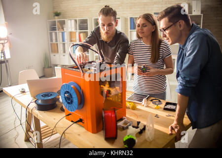 Three young engineers standing at wooden desk and using 3D printer in order make small prototypes, interior of modern laboratory on background Stock Photo