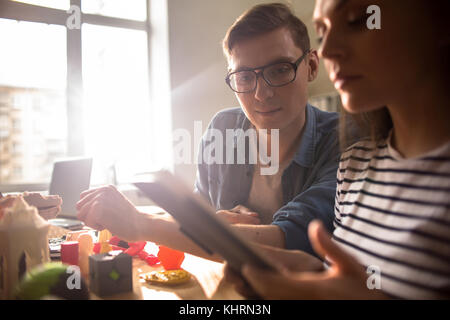 Profile view of pretty young engineer using digital tablet while sitting at desk next to male colleague and thinking over joint project, lens flare Stock Photo