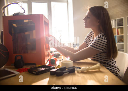 Profile view of beautiful young engineer wearing striped T-shirt sitting at office desk and using 3D printer, lens flare Stock Photo