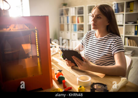Waist-up portrait of confident young designer sitting at desk with digital tablet in hands and keeping eye out for process of 3D printing Stock Photo