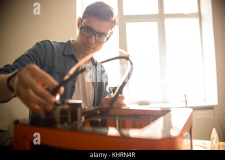 Waist-up portrait of concentrated young technician in eyeglasses repairing 3D printer after breakage, interior of spacious laboratory on background Stock Photo